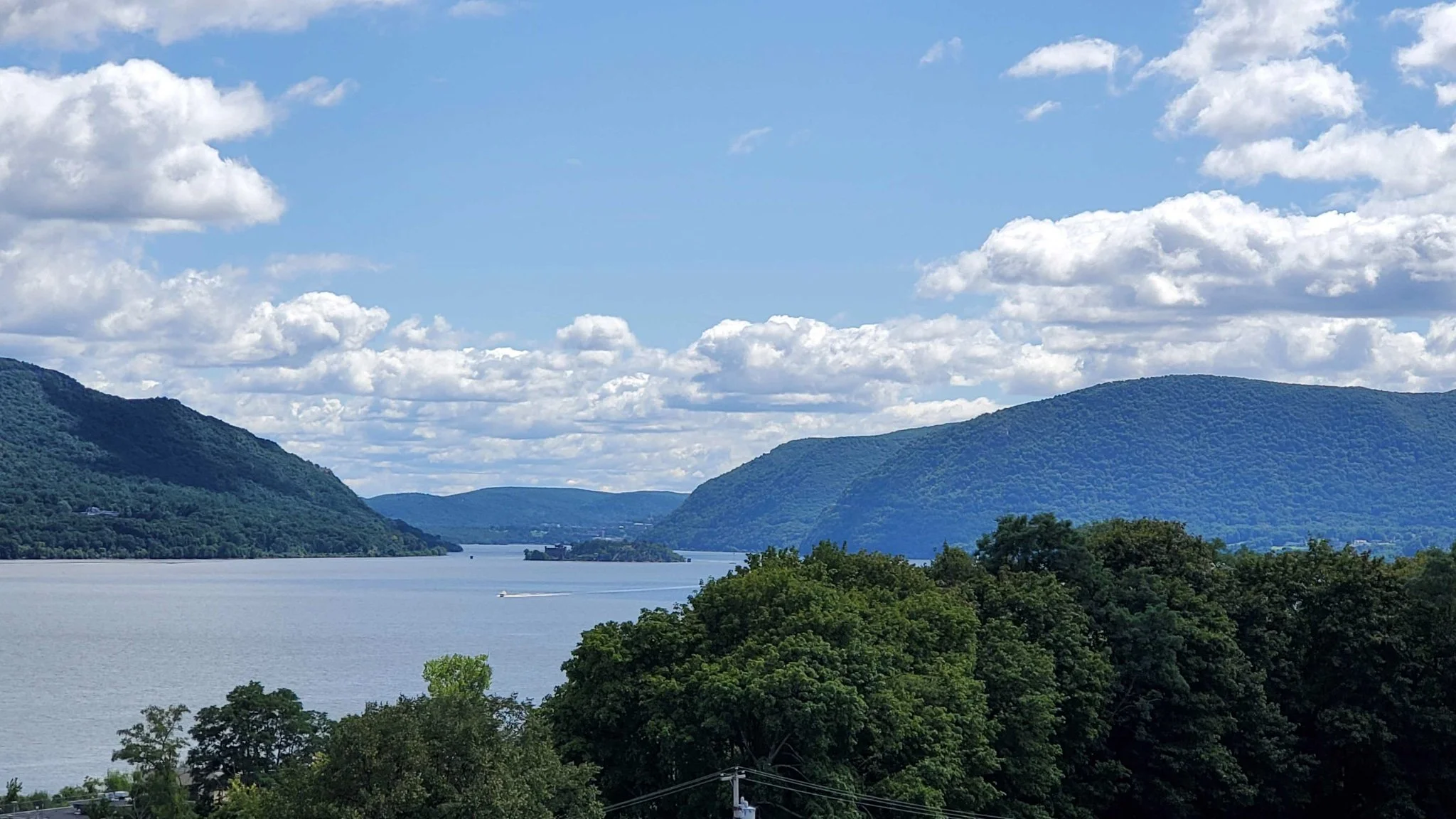 A scenic view of a river surrounded by green hills and mountains under a partly cloudy sky with scattered white clouds.