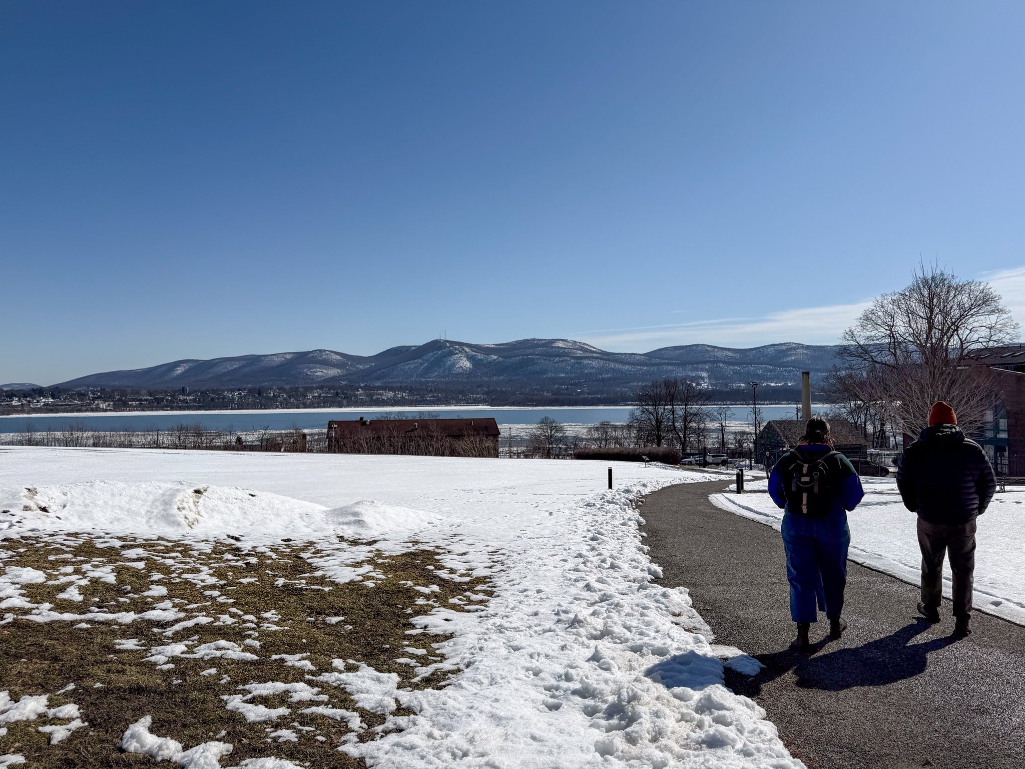 Two people walking on a paved path in a snowy landscape with mountains in the background under a clear blue sky.