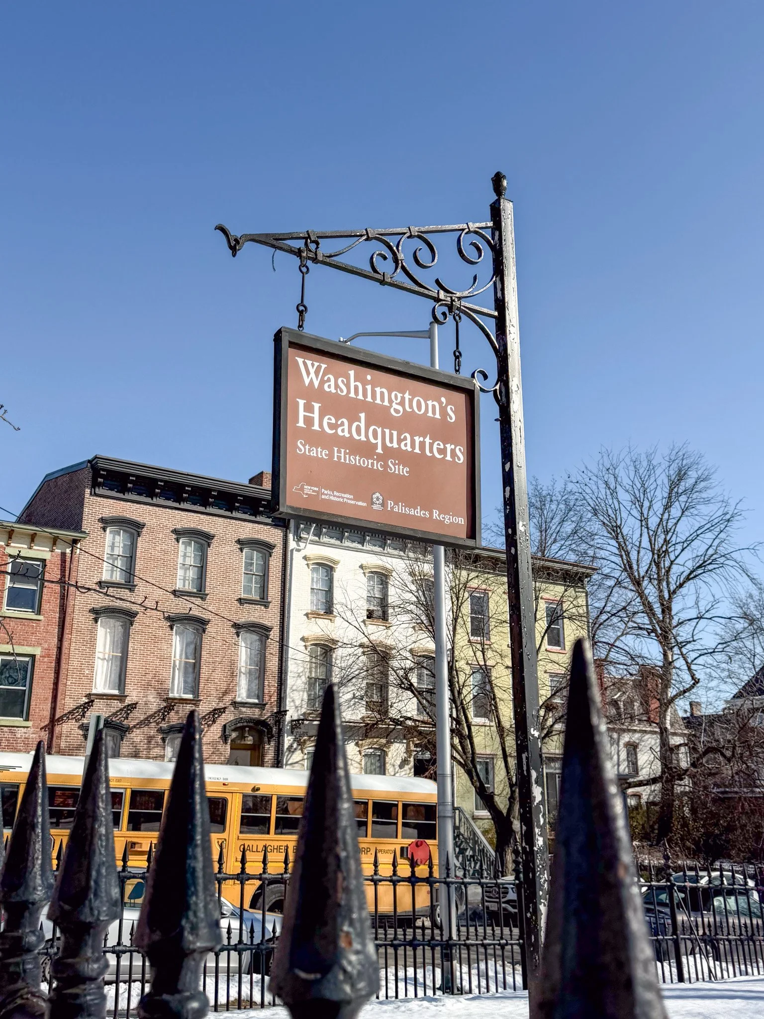 A sign reading 'Washington's Headquarters State Historic Site' hanging from an ornate black metal post, with a brick building, trees, a yellow school bus, and a black iron fence in the background under a clear blue sky.