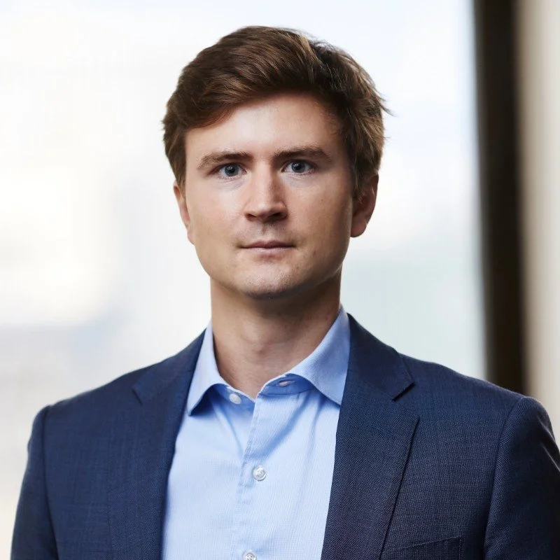 A photo of Max Mortimer, who has brown hair, wearing a navy suit with a sky blue shirt.