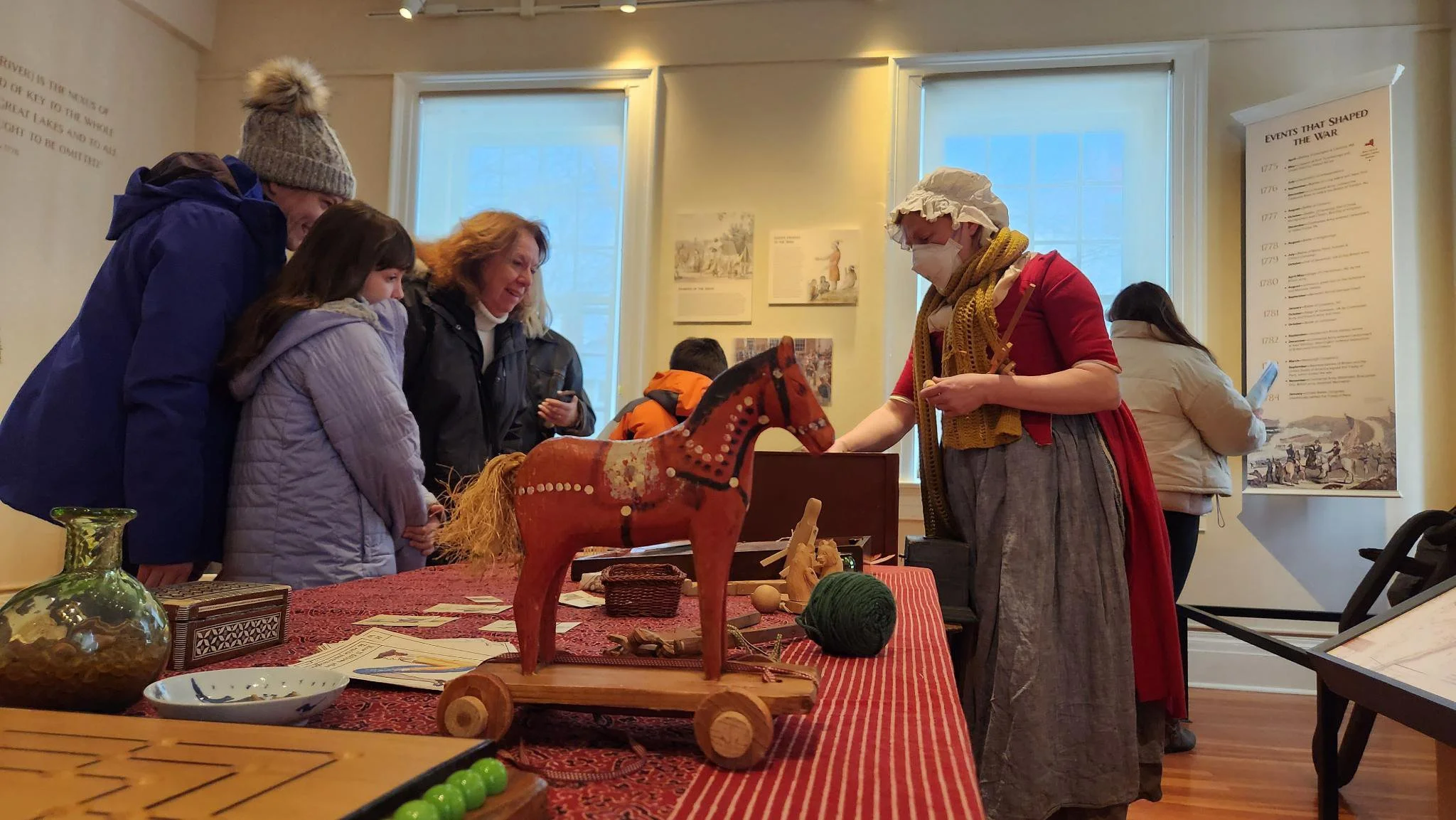 A woman in historical costume with a bonnet and face mask demonstrating a wooden toy horse at a museum or historical exhibit while a group of children and adults observes.
