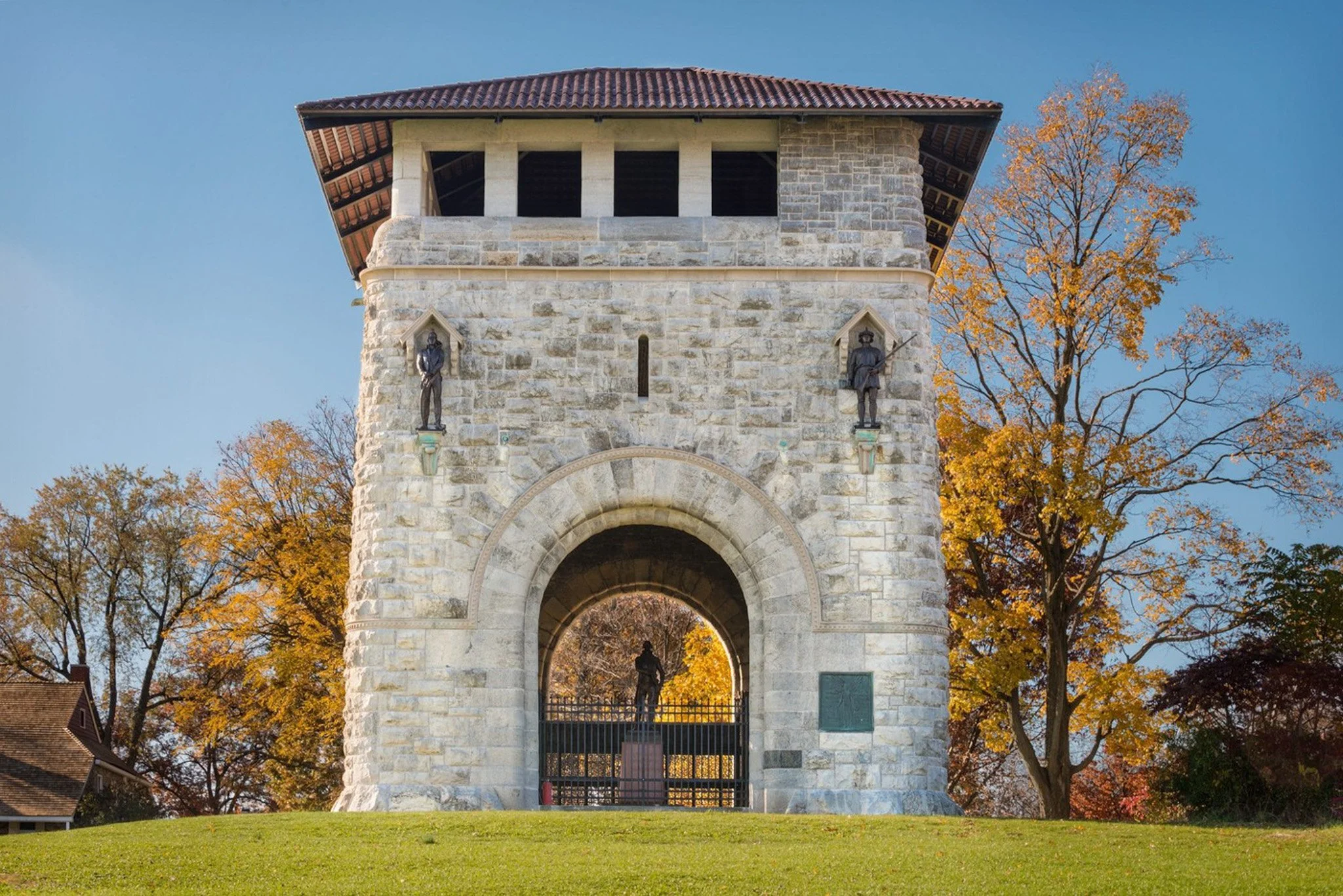 Stone tower with arched gate and statues of historical figures, surrounded by fall-colored trees on a grassy hill.