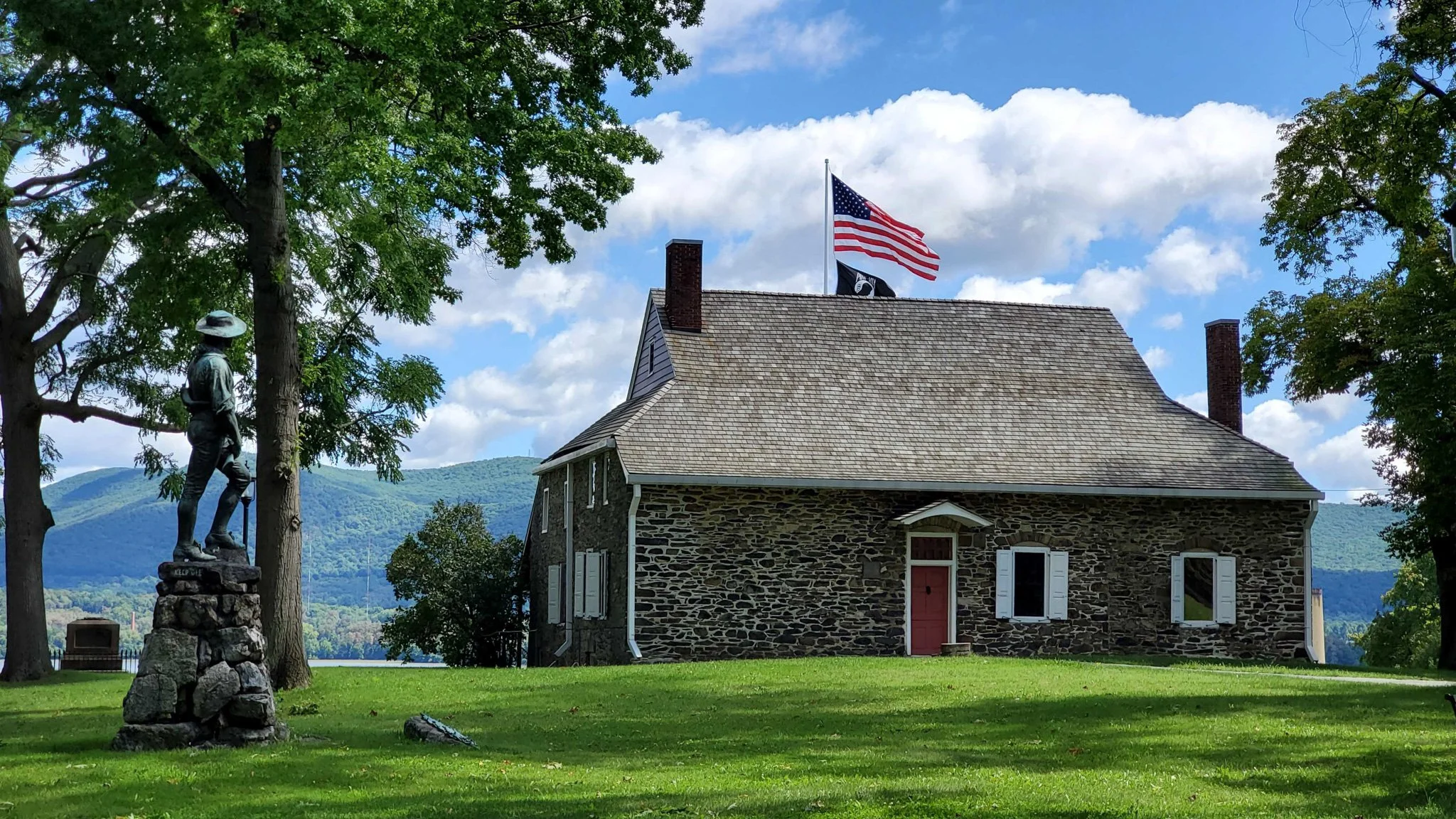 A stone house with white shutters and a red door, surrounded by green grass and trees. An American flag and another black flag with a white emblem are flying on the roof. A mountain and body of water are visible in the background.