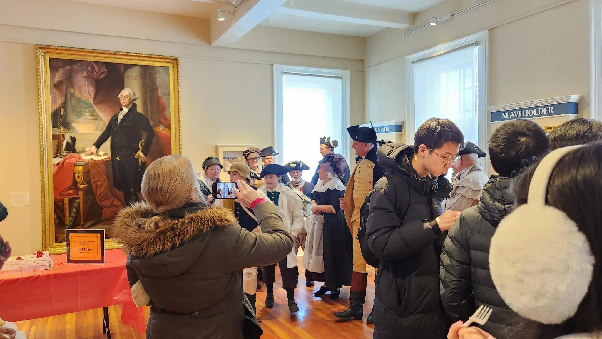 Visitors in colonial-era costumes gather in an art museum, taking photos and viewing displays, with a large portrait of George Washington on the wall.