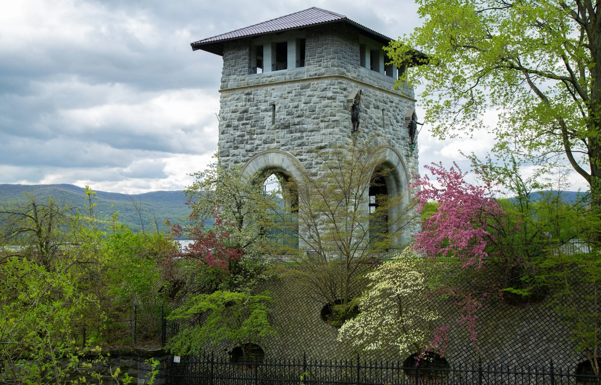 A stone tower with an arched opening and small windows, surrounded by flowering trees and greenery, with mountains in the background under a cloudy sky.