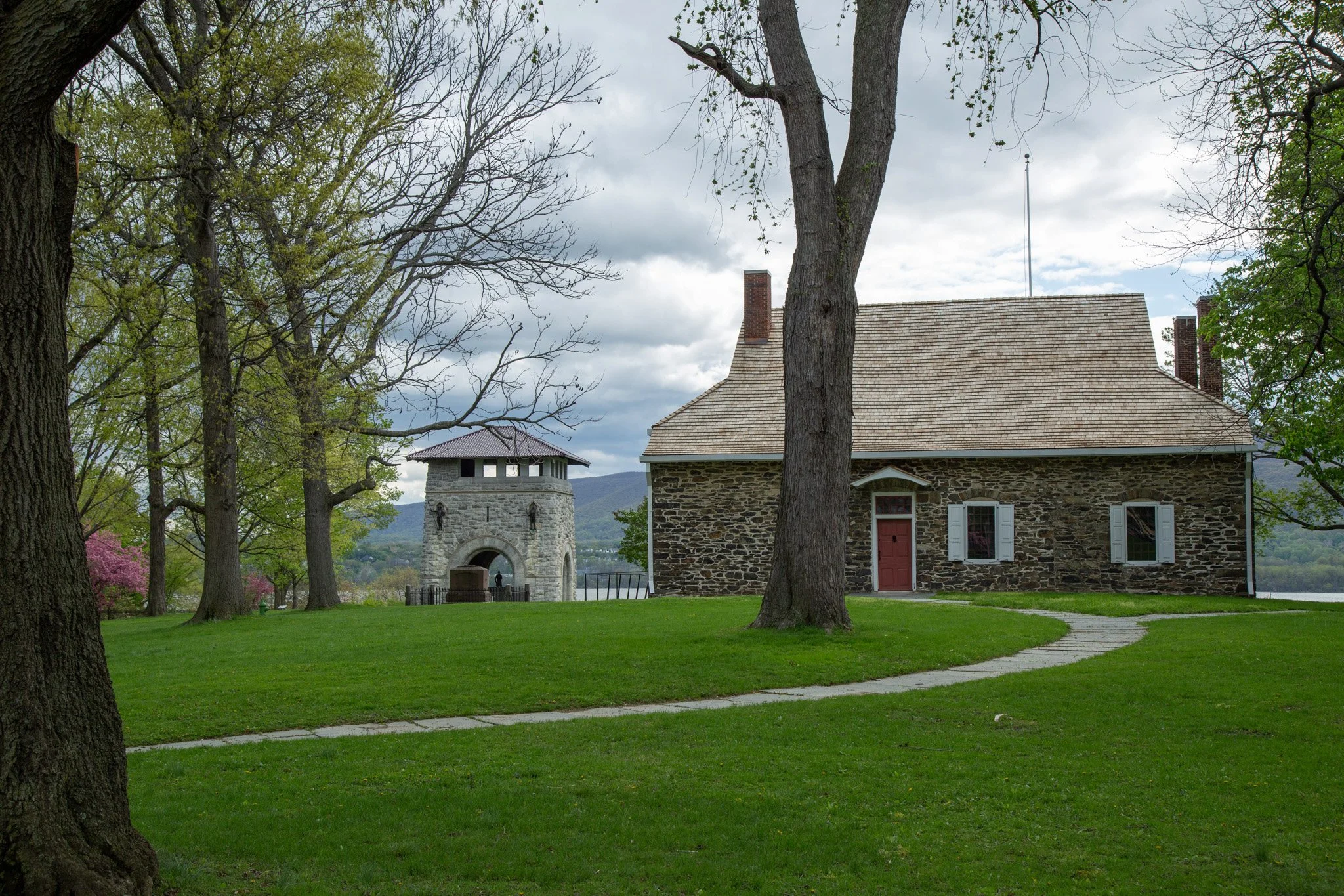 A stone house with a red door and white window shutters on a grassy lawn surrounded by large trees, with a stone tower in the background along a body of water under a cloudy sky.