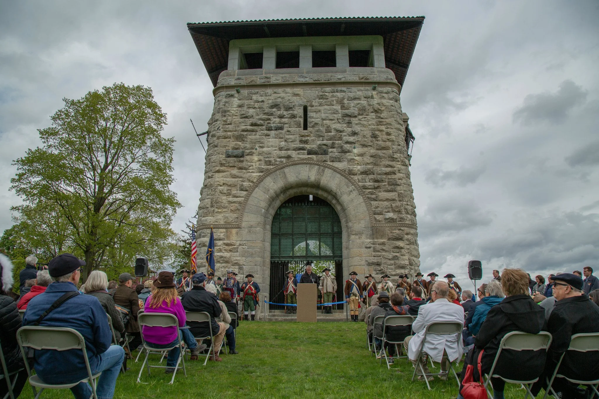 A large gathering of people attending an outdoor event in front of a historic stone tower, with some individuals dressed in colonial military uniforms, and flags displayed.