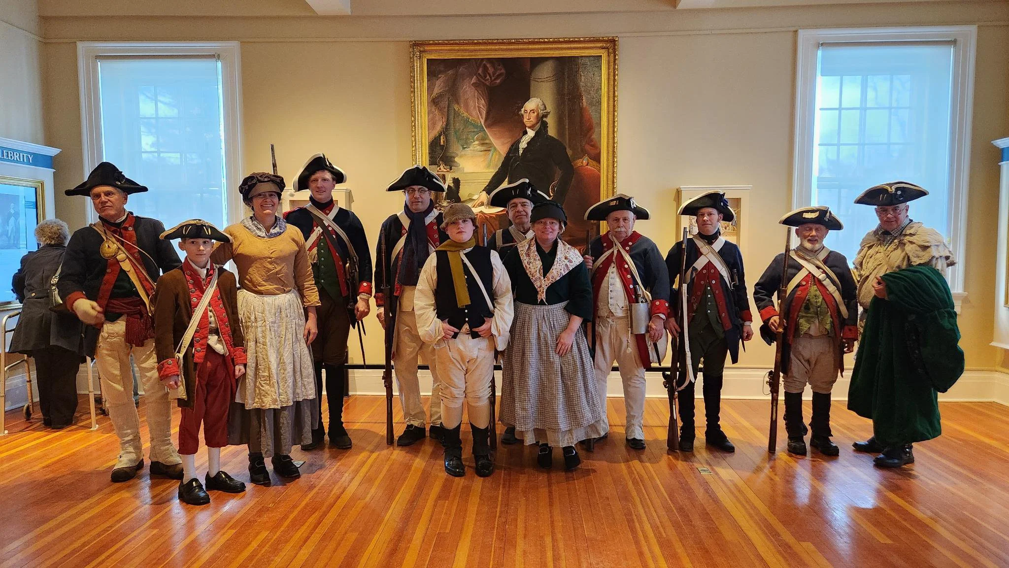 Group of people dressed in Colonial American military costumes posed in a museum with a portrait of George Washington in the background.