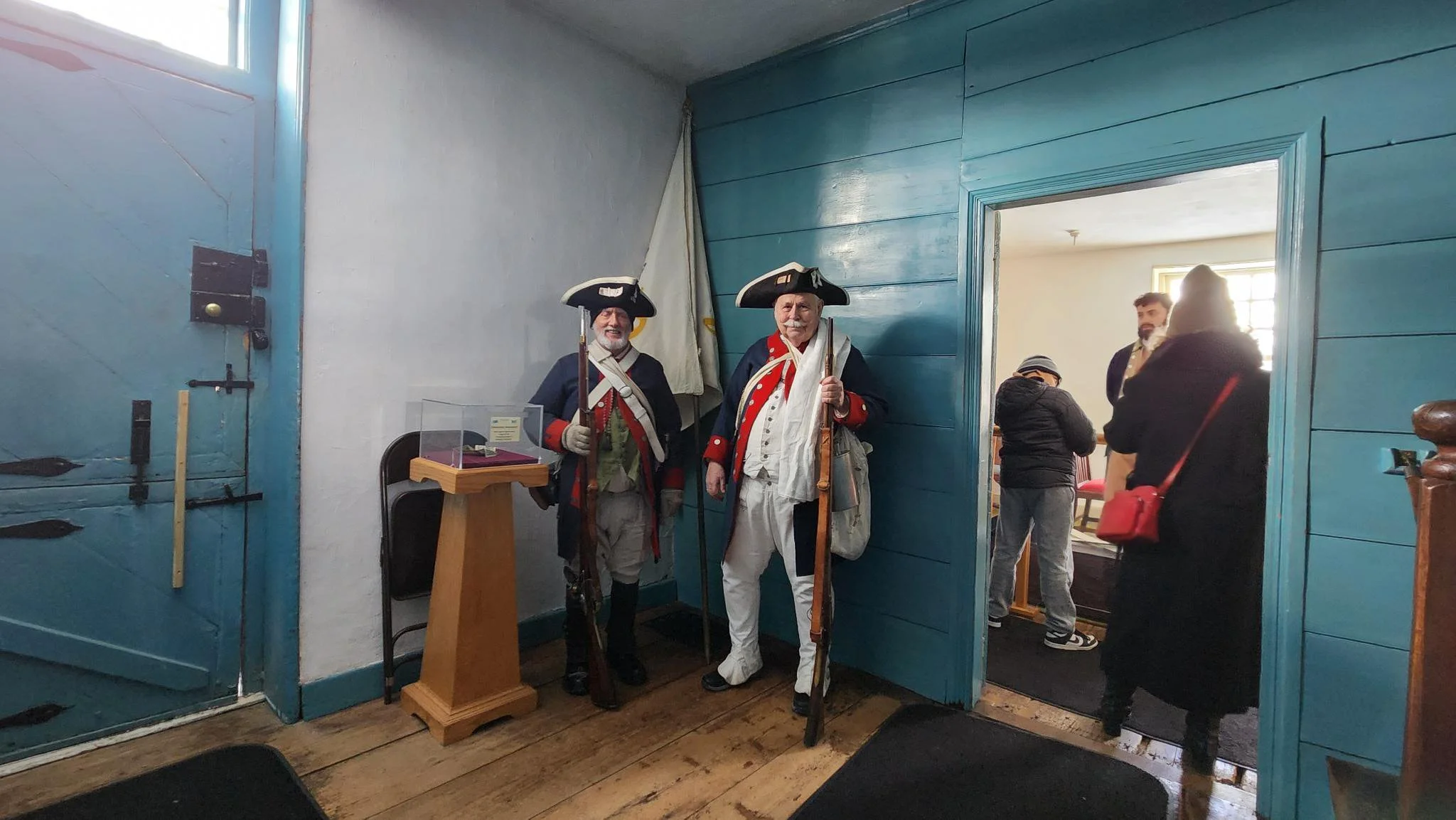Two men dressed as historical soldiers in uniforms with tricorn hats, standing near a blue wall and doorway inside a building, with other people visible through the doorway.