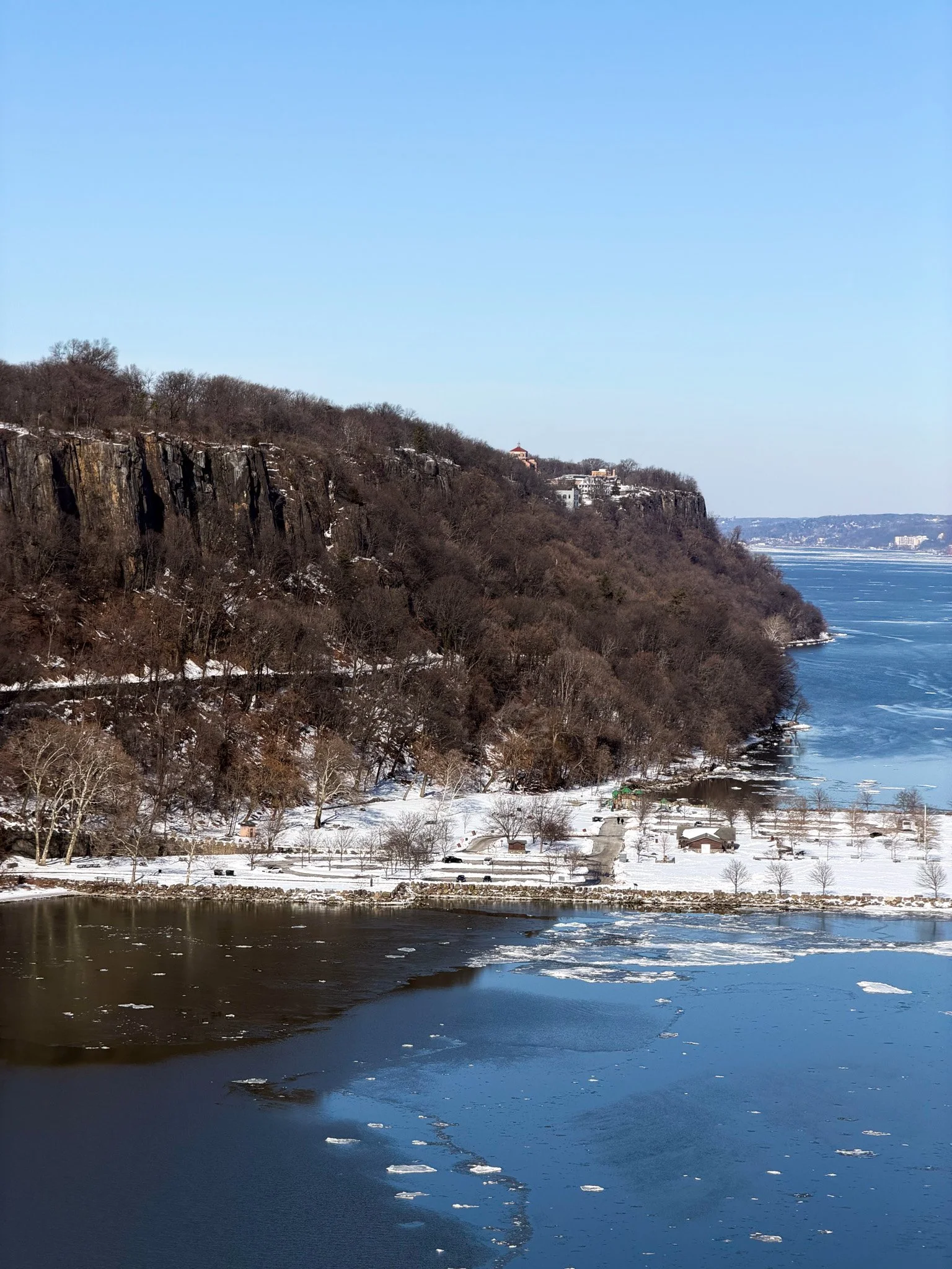 A winter landscape with a river partially frozen, snow-covered land, leafless trees, a hillside with cliffs, and buildings in the distance under a clear blue sky.