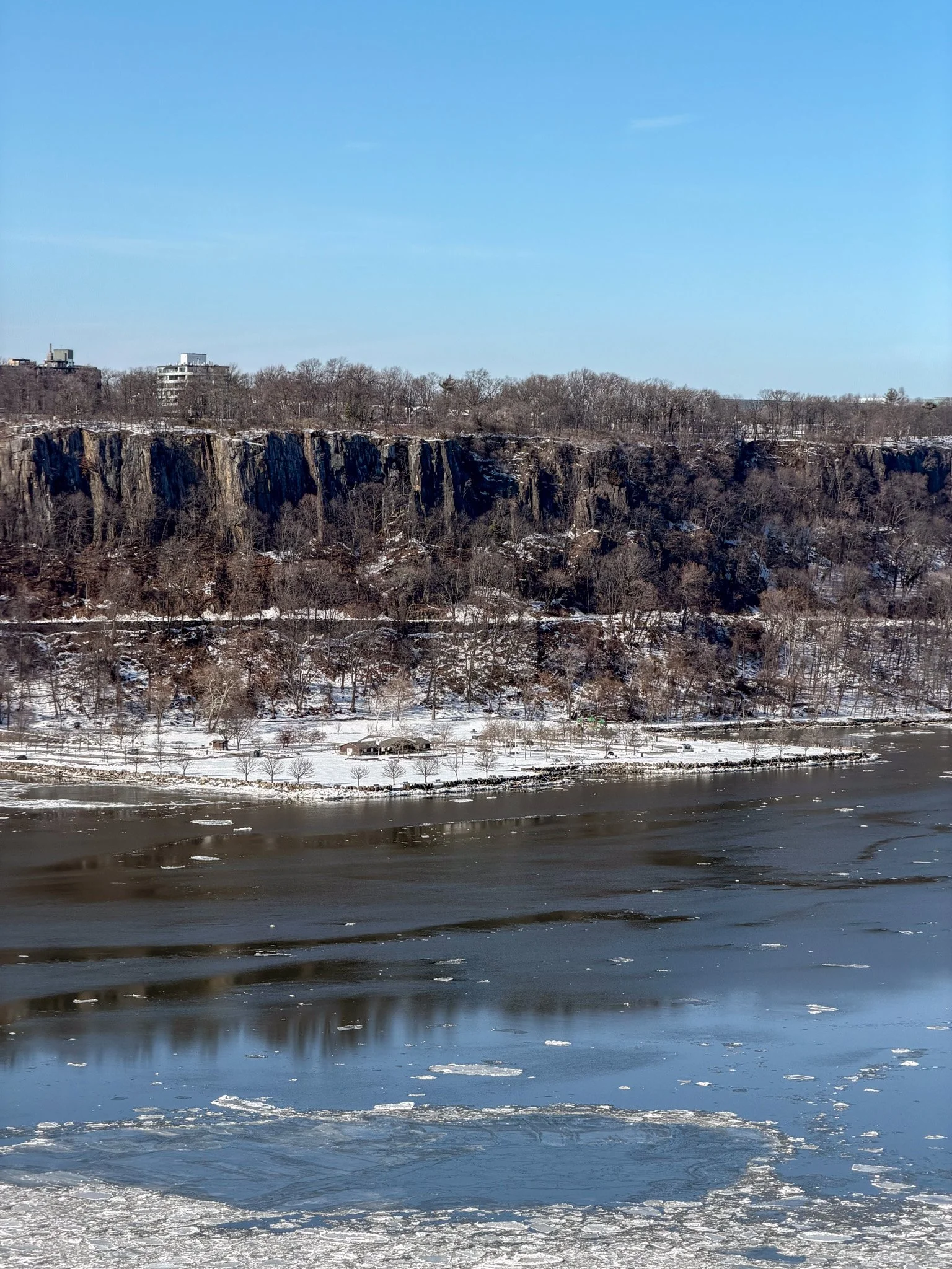 A winter landscape with a river partially frozen, snow-covered trees along the riverbank, a cliff with bare trees and some buildings in the background under a clear blue sky.