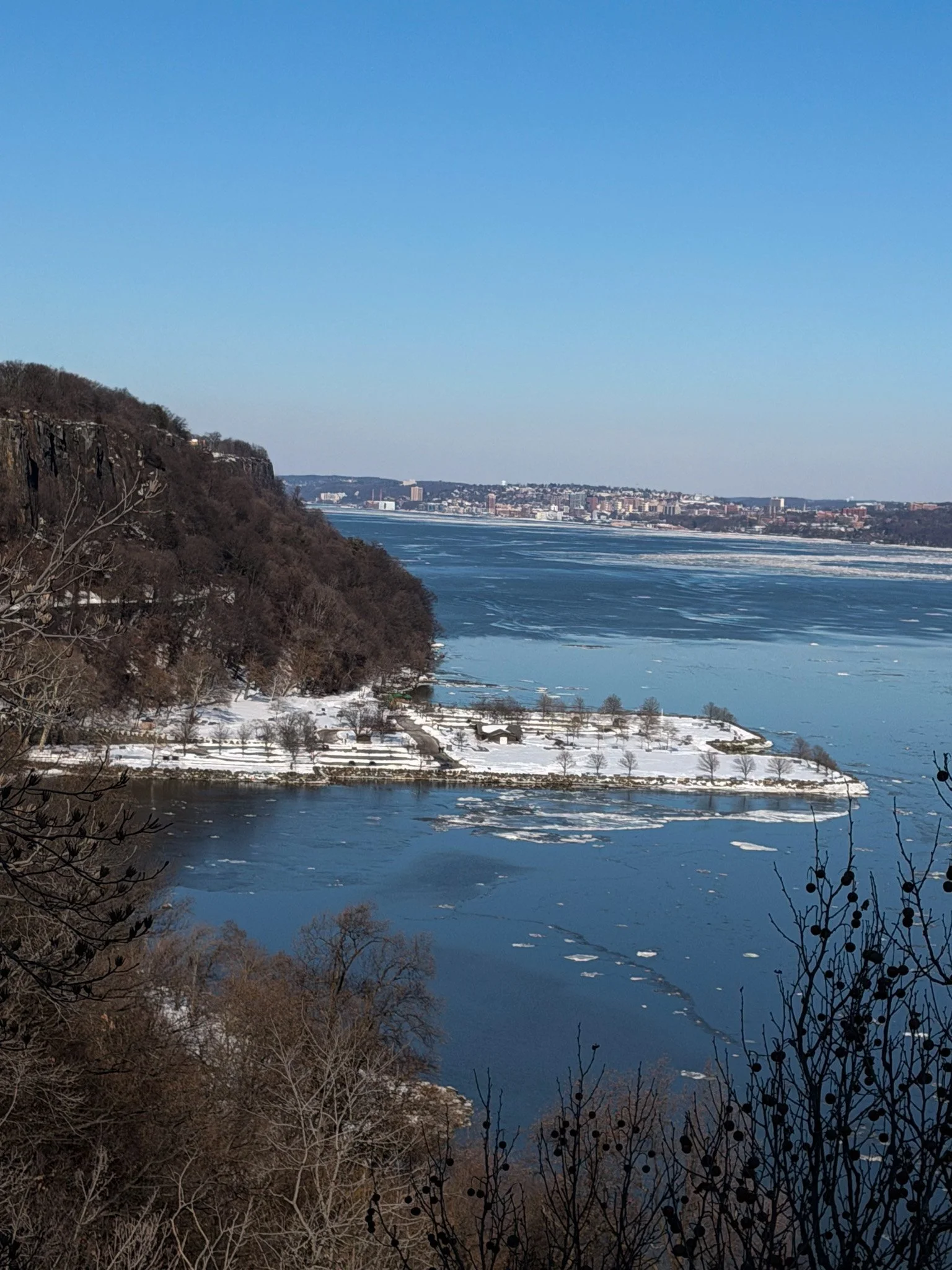 A scenic winter landscape of a river with ice, snow-covered banks, and leafless trees, with a city skyline in the background under a clear blue sky.
