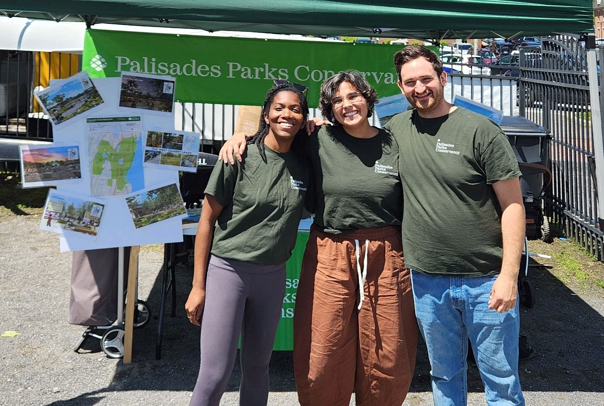 Three people wearing olive green Palisades Parks Conservancy tshirts pose together in front of a tabling booth.