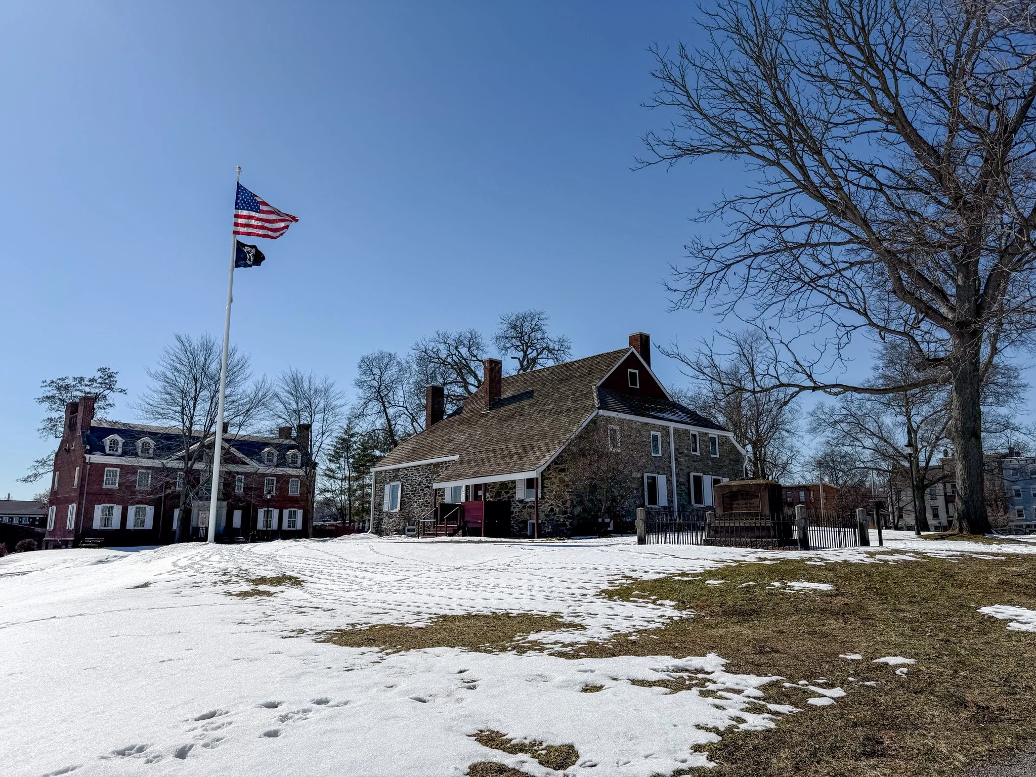 A historic stone building with a steep roof, surrounded by leafless trees, on a partly snow-covered lawn under a clear blue sky. An American flag and a black flag are flying on a tall flagpole nearby.