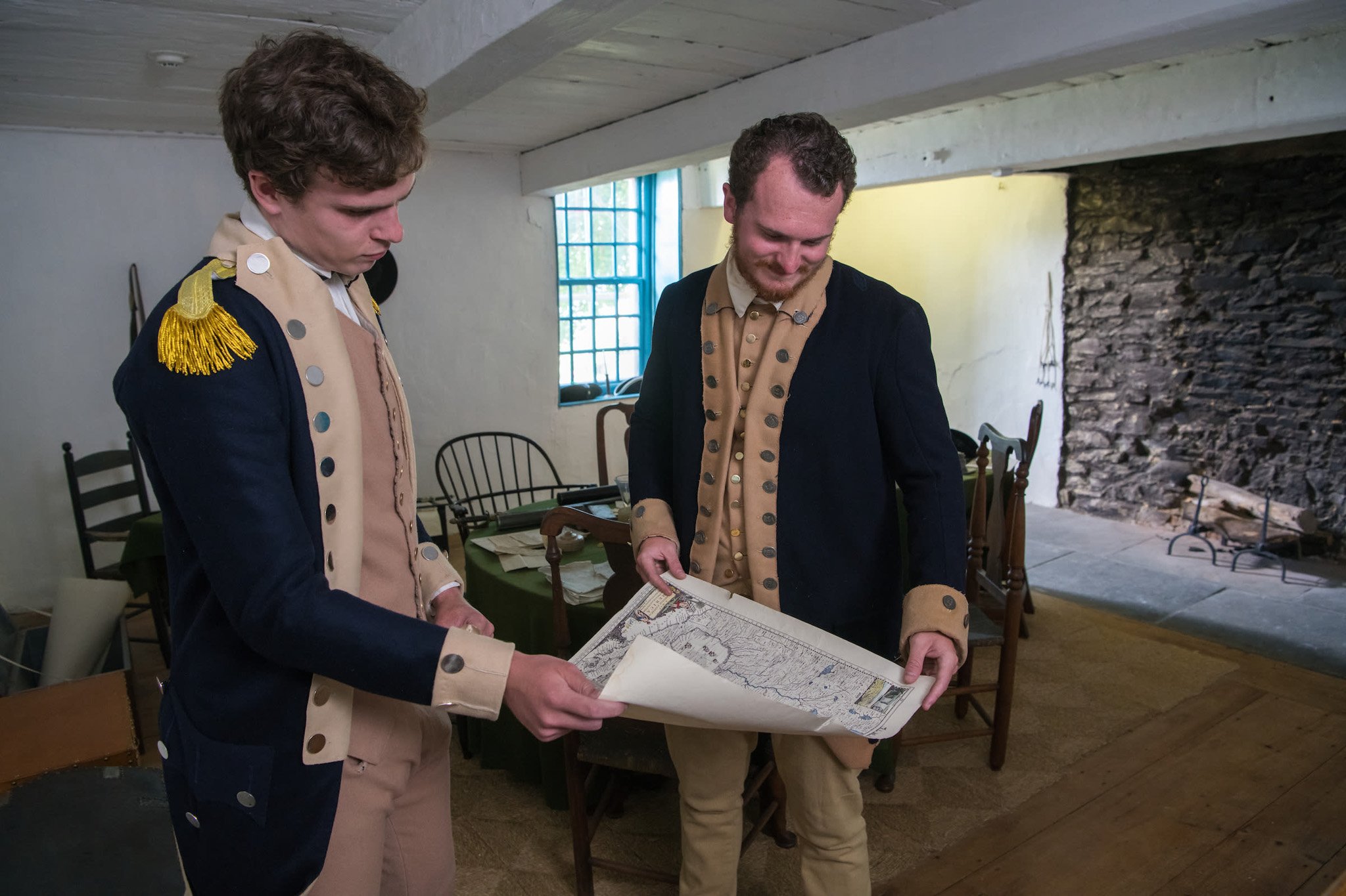 Two men in historical military costumes examining a map indoors, with a stone fireplace and wooden furniture in the background.