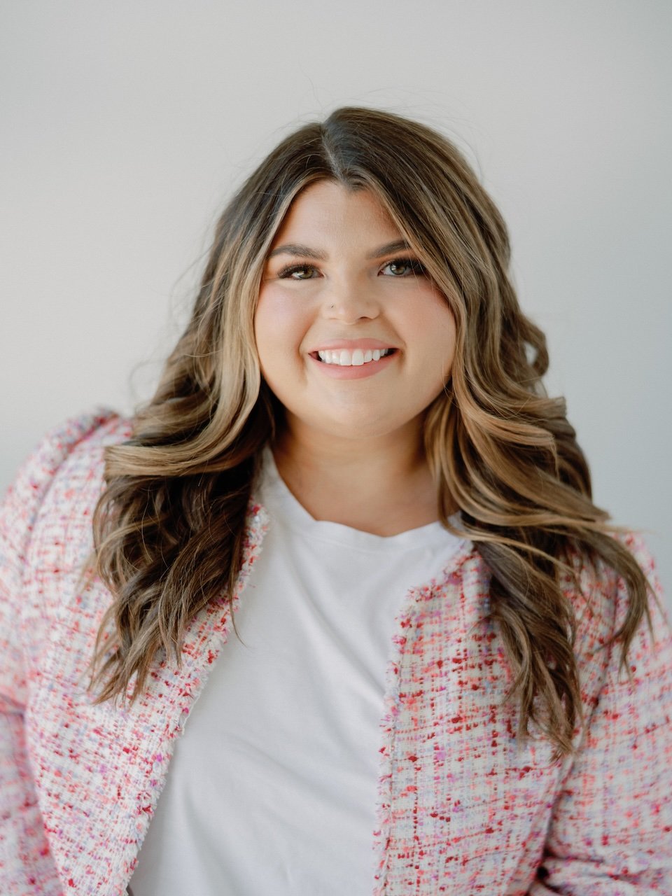 Portrait of a young woman with wavy brown hair, smiling, wearing a white top and pink patterned blazer against a plain background.