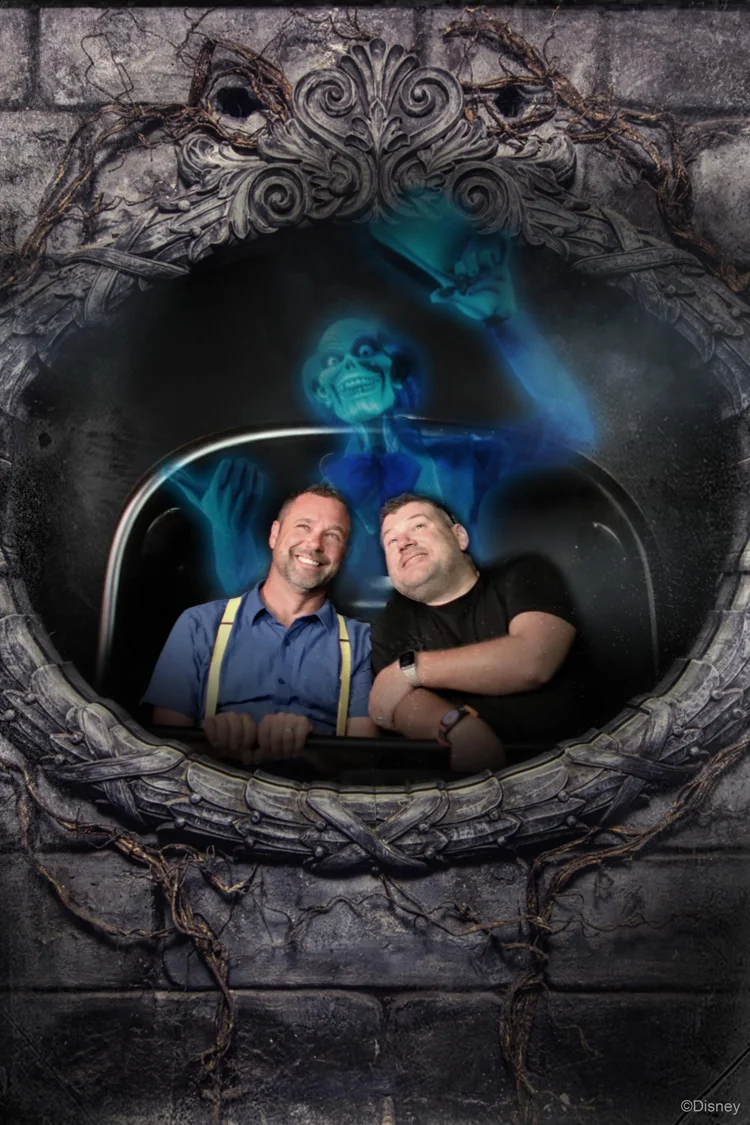 Two men smiling and posing inside a themed photo booth with a spooky mirror and ghostly figure in the background, surrounded by a darker, eerie frame with vines.
