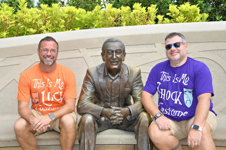 Two smiling men sitting on either side of a bronze statue of a seated man, outside on a bench with a lush green background. Both men are wearing colorful t-shirts and sunglasses.