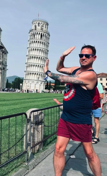 A man with sunglasses and tattoos standing in front of the Leaning Tower of Pisa, making a pose with his hand against the tower.