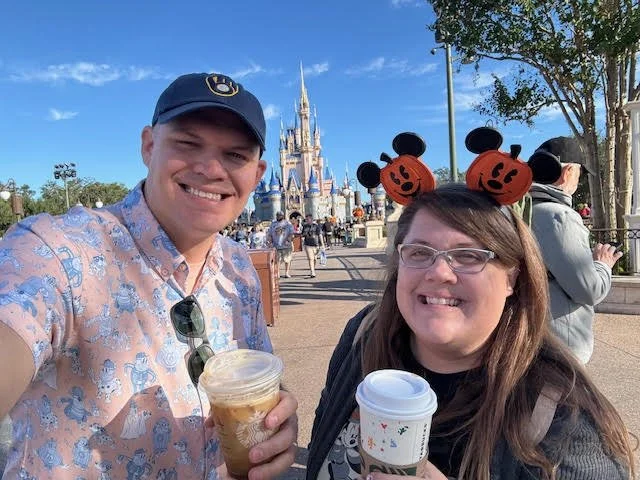 Two smiling people at Disney park, with Cinderella Castle in the background. The man is wearing a blue cap and patterned shirt, holding a cup of iced coffee. The woman is wearing Mickey Mouse headbands with pumpkin faces, glasses, and a dark top, holding a hot beverage. Other park visitors and trees are visible.