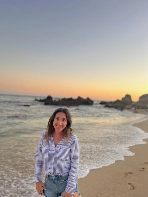 A woman smiling on a beach at sunset, with ocean waves and rocks in the background.