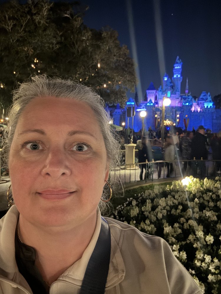 A woman taking a selfie at Disneyland at night with Sleeping Beauty Castle illuminated in the background.