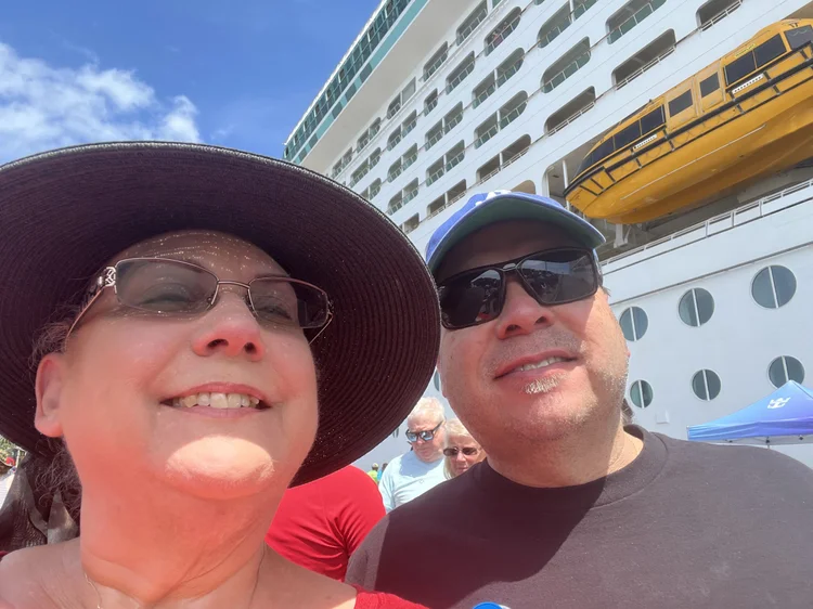 Two people taking a selfie on a cruise ship with the ship's exterior, windows, and yellow lifeboat visible in the background. The woman is wearing a large sunhat and glasses, while the man is wearing a baseball cap and sunglasses.