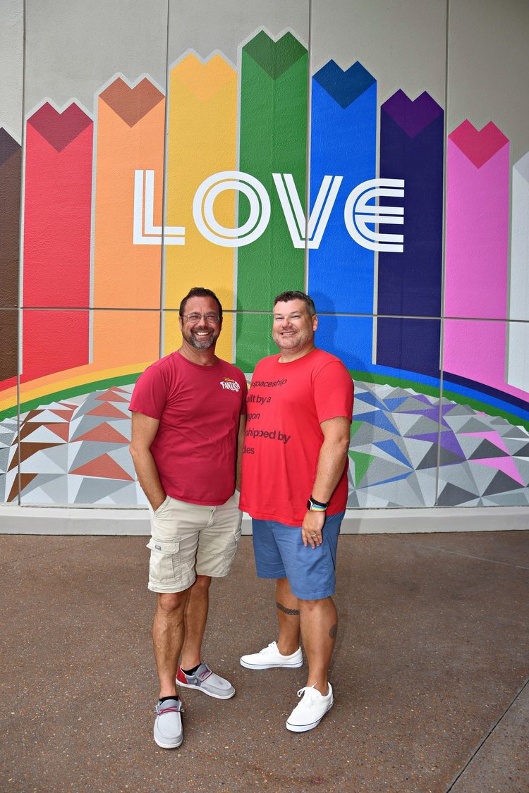 Two men standing in front of a colorful rainbow mural with the word 'LOVE' written across it, smiling at the camera.