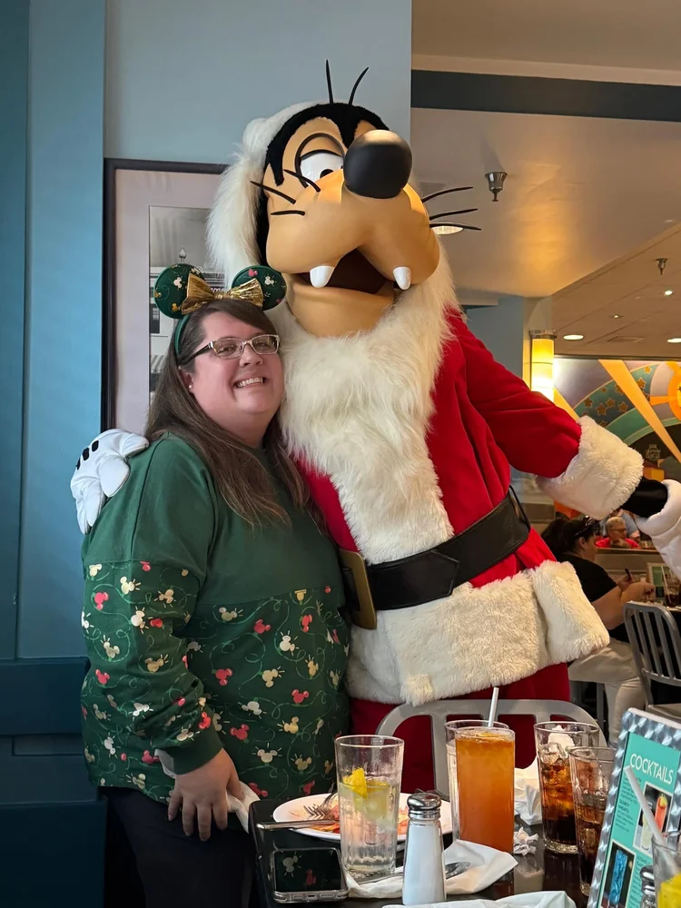 A woman smiling in front of a large costumed character of Goofy dressed as Santa Claus at a restaurant during Christmas.