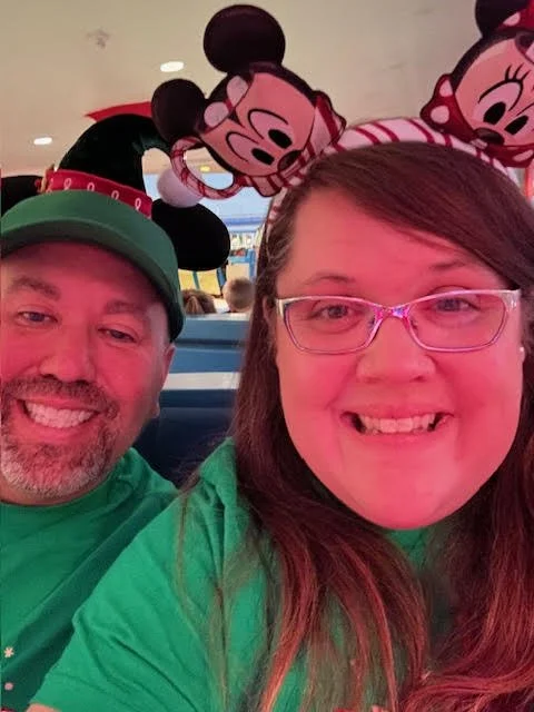 A smiling man and woman wearing green shirts, with the man in a Christmas hat and the woman wearing glasses, in front of Minnie Mouse balloons at a Disney theme park.