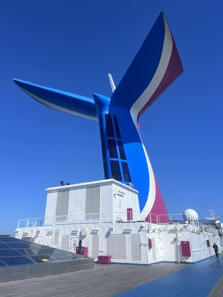 Photo of a large ship's funnel painted in blue, white, and red, resembling an abstract whale tail, against a clear blue sky.