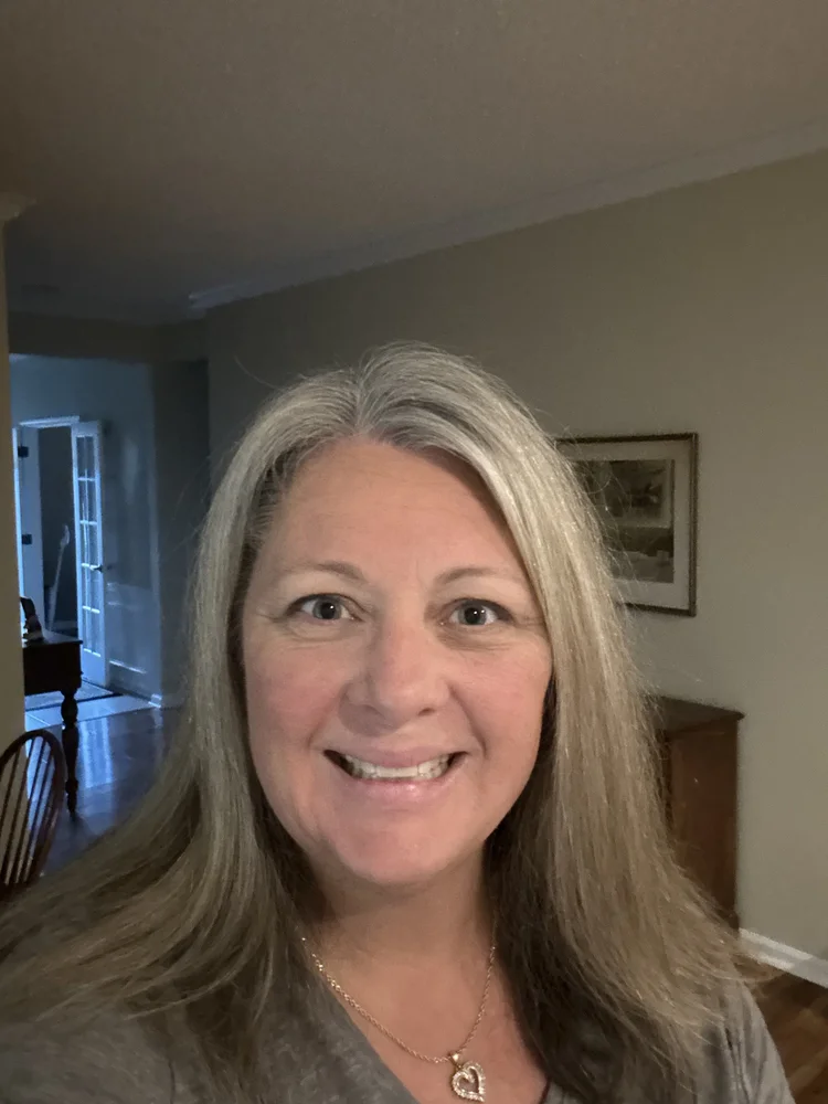 Close-up of a smiling woman with long gray hair, wearing a gray top and a heart-shaped pendant necklace, in a room with a framed picture on the wall and a wooden cabinet in the background.