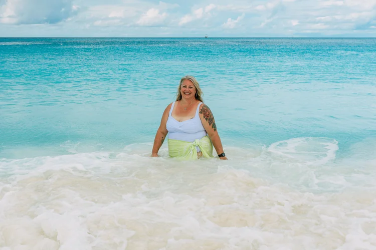 A woman standing in the ocean with waves around her, smiling, with a light green sarong and white top, tattoos on her arms, and a smartwatch on her wrist.
