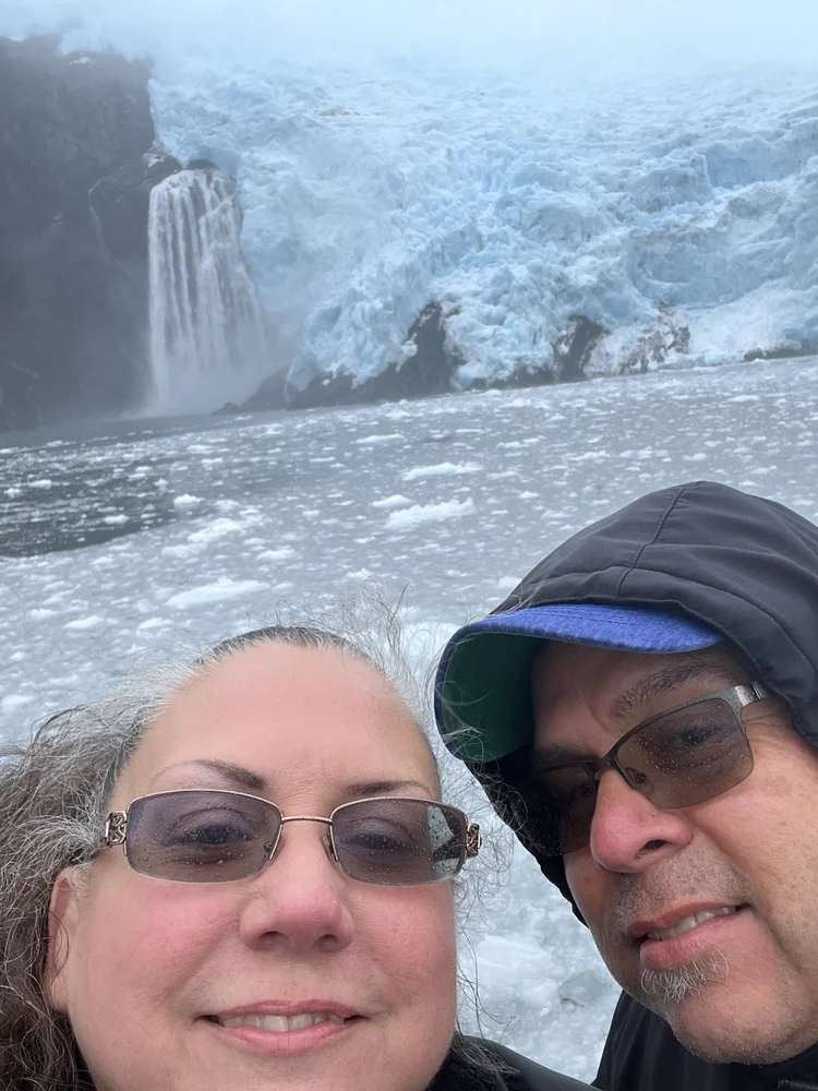 A smiling couple taking a selfie in front of a large icy waterfall with ice and snow in the background.