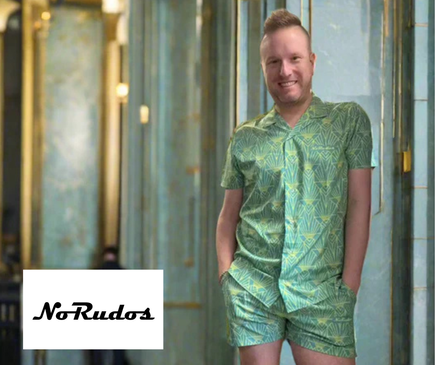 A young man with short, styled hair wearing a matching green patterned short-sleeve shirt and shorts, standing against a blue tiled wall with a reflection in a window behind him, smiling at the camera, with a white rectangular logo reading NoRudos in the lower left corner.