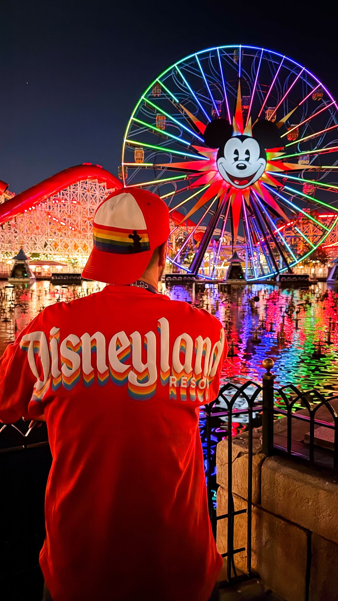 A traveler in a rainbow Disneyland Resort jacket and Pride cap watching the Pixar Pal-A-Round wheel light up at night