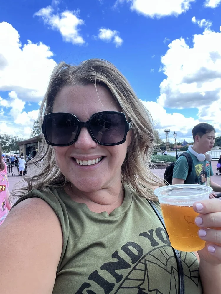 A smiling woman with blonde hair and large sunglasses holding a cup of beer at a theme park.
