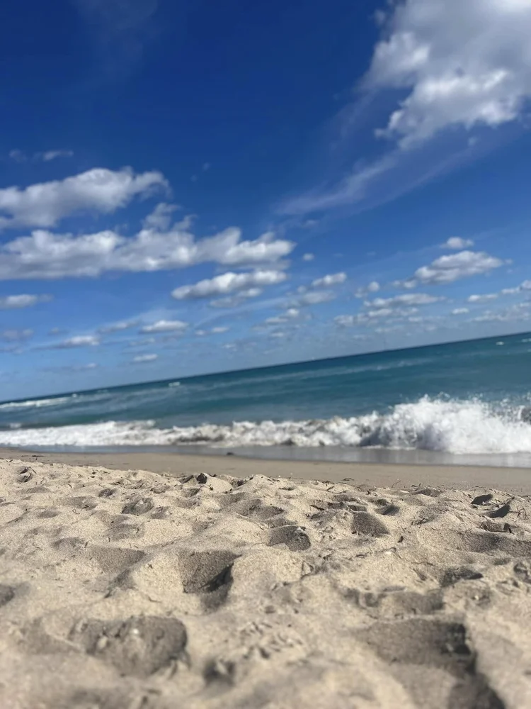 Sandy beach with footprints, ocean waves, and a blue sky with scattered clouds.