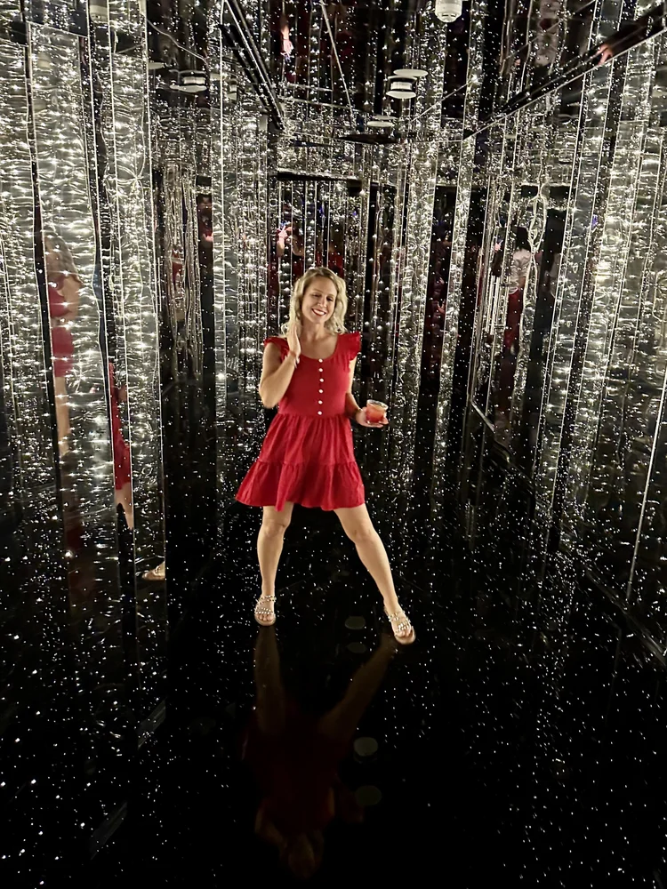 A woman in a red dress smiling and posing inside a mirrored, starry-themed room, holding a drink.