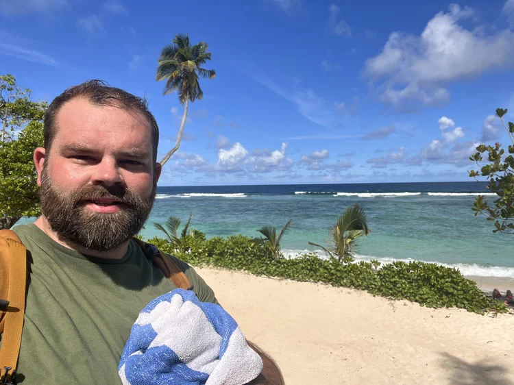 A man with a beard and short hair taking a selfie at a beach with blue sky, palm trees, ocean, and sandy shore in the background.