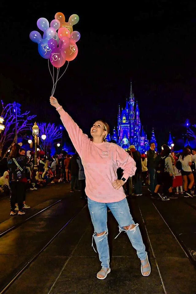 A woman holding colorful balloons at Disneyland at night with the castle illuminated in the background.