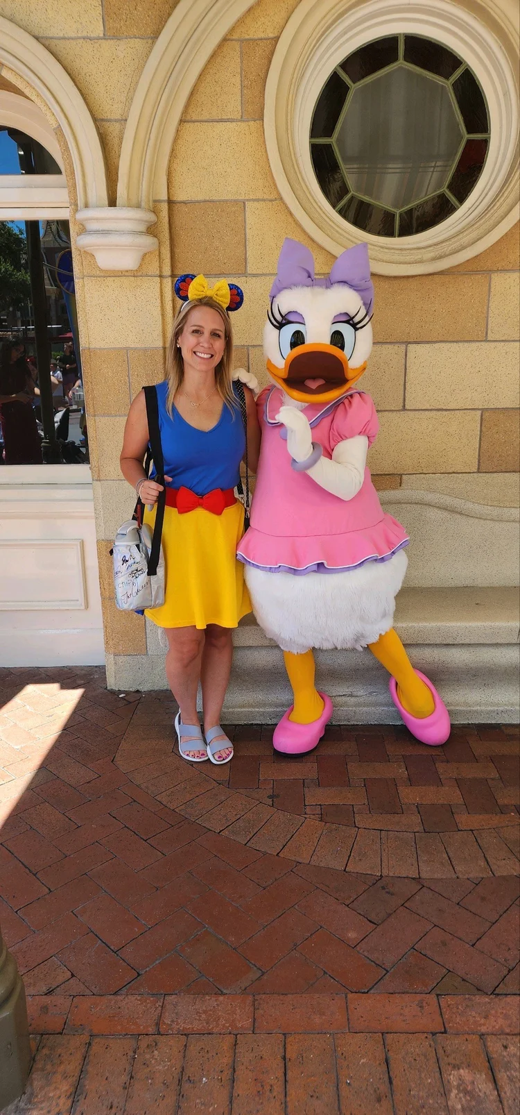 A woman stands next to a person dressed as Daisy Duck at a theme park, both smiling. The woman wears a blue top, a yellow skirt with a red bow, and Minnie Mouse ears. The background features a brick sidewalk and a castle-like building.