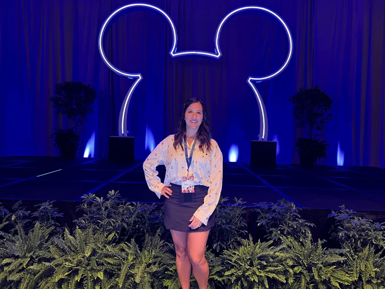 A woman standing on a stage with a large illuminated Mickey Mouse head outline in the background at a convention or event.