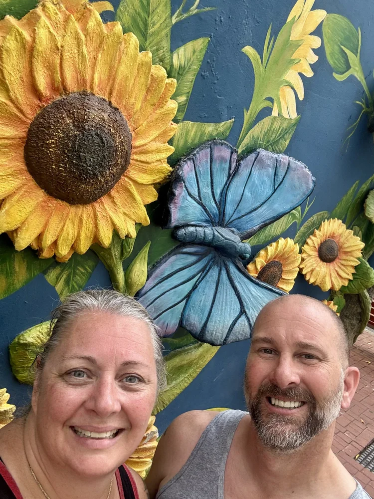A smiling woman and man taking a selfie in front of a colorful wall mural featuring sunflowers and a blue butterfly.