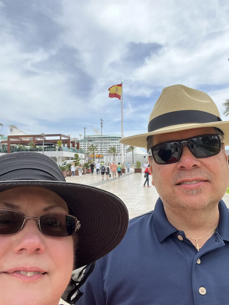 A man and woman wearing hats and sunglasses taking a selfie outdoors with a cruise ship, a Spanish flag, palm trees, and people walking in the background.
