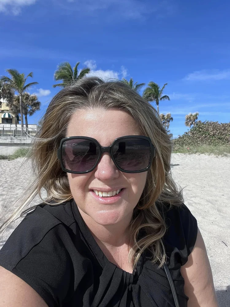 Woman with wavy blonde hair wearing large black sunglasses smiling at the beach with sand, palm trees, and buildings in the background under a blue sky.