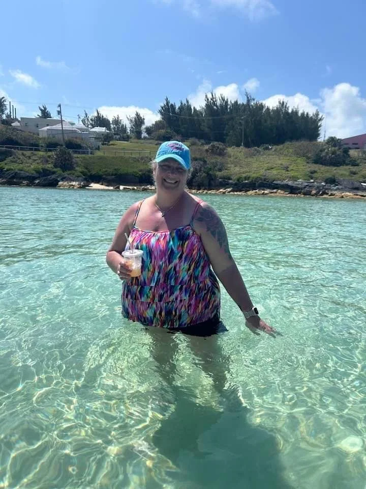 A woman standing in clear, shallow water at the beach, smiling and holding a drink, wearing a colorful tank top, a blue cap, and a watch, with a backdrop of trees, houses, and a partly cloudy sky.