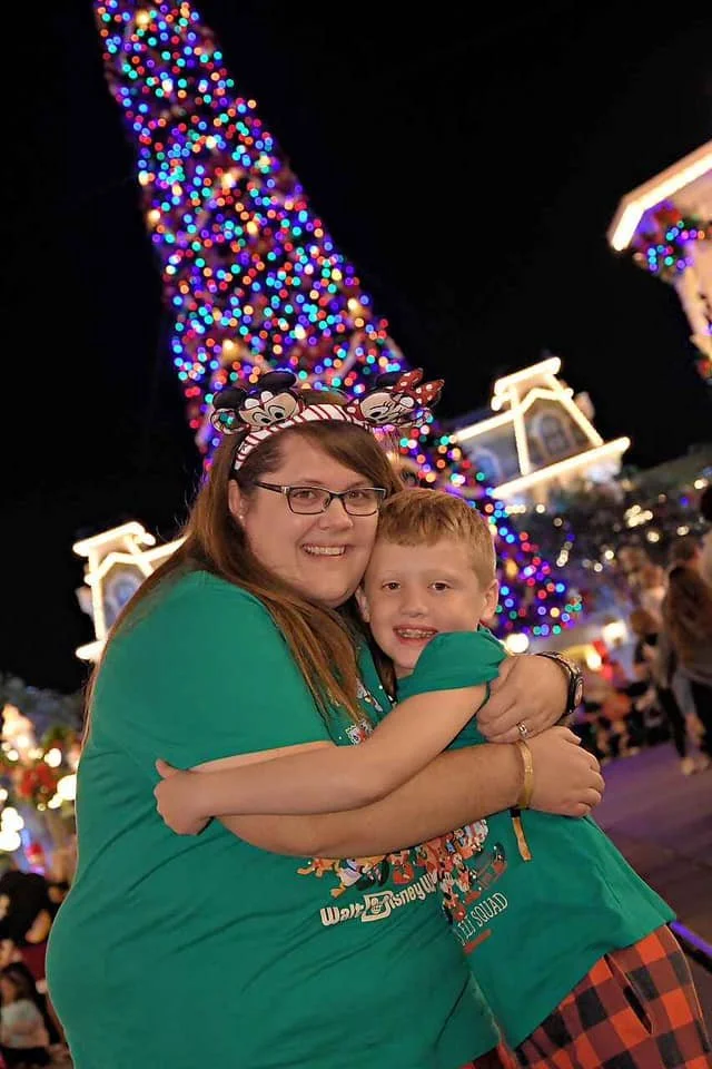 A woman and a young boy hugging in front of a large, decorated Christmas tree with colorful lights, with buildings and other people in the background at night.