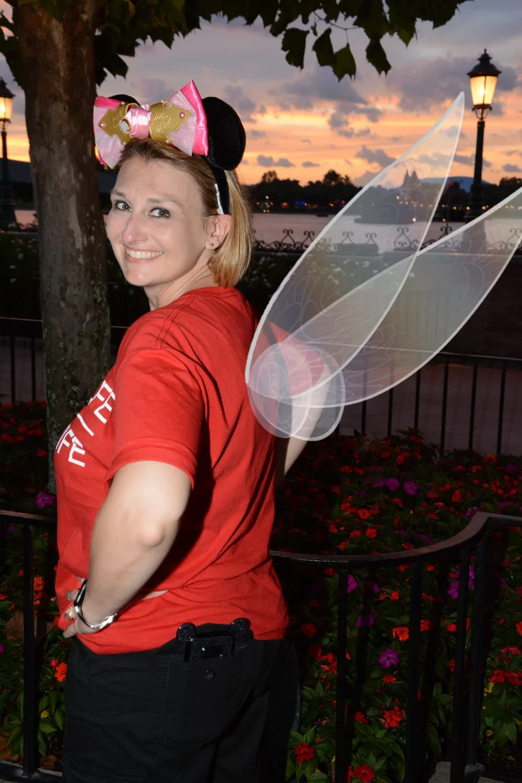 A woman smiling at sunset, wearing a red shirt, a pink and gold Minnie Mouse headband, and fairy wings, standing outdoors near a tree, with flowers and a body of water in the background.