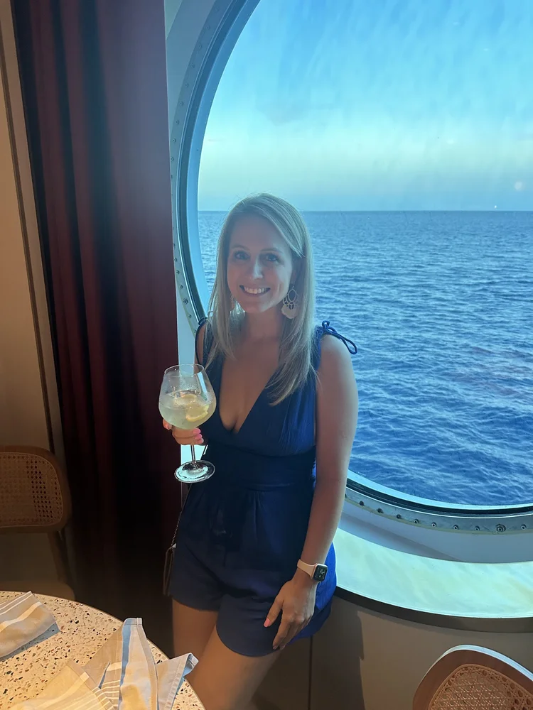 Woman in blue dress holding a glass of white wine or cocktail, standing in front of a large round window looking out onto the ocean, on a cruise or ship.